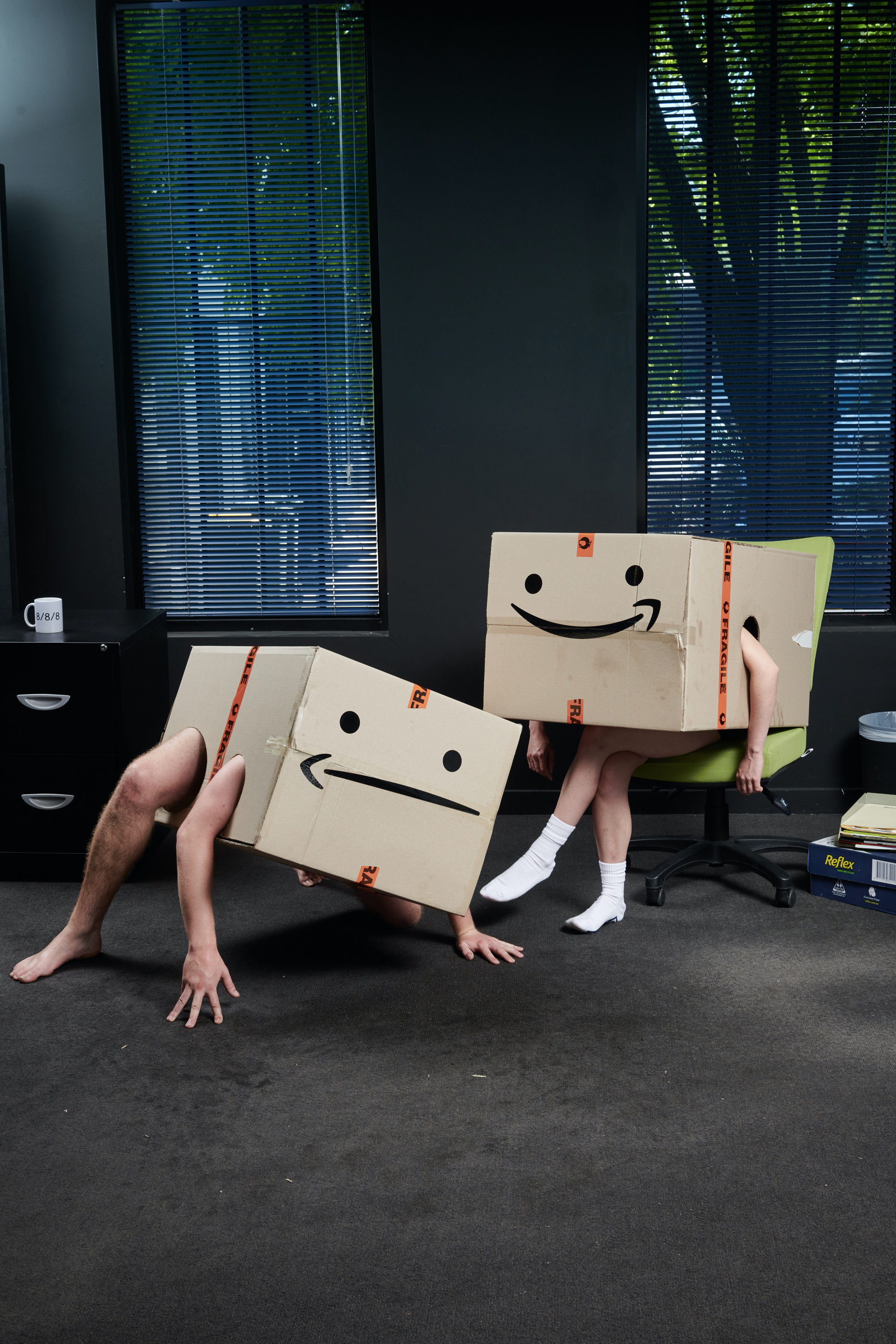 A man and a woman wear cardboard boxes with holes for only their arms and legs. He is crouched on the ground, she is seated.