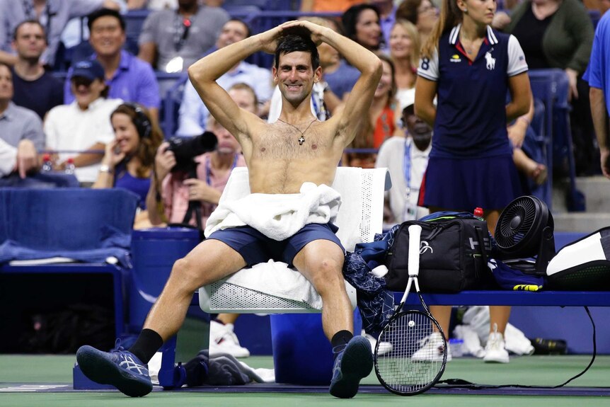 A shirtless Novak Djokovic rests his hands on his head, as he smiles sitting by the tennis court