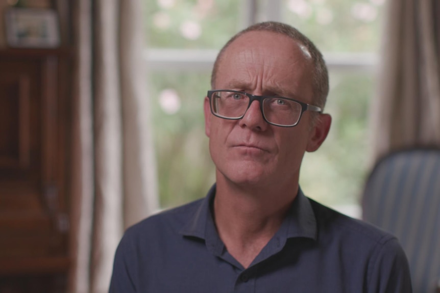 A middle-aged man with short hair and glasses looks into the camera with a serious expression.