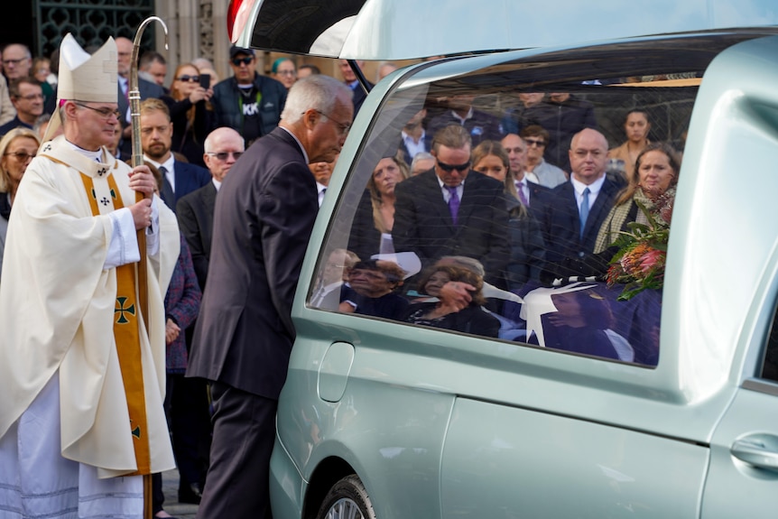 Mourners bow their heads in the background as a funeral director attends to the coffin in the hearse