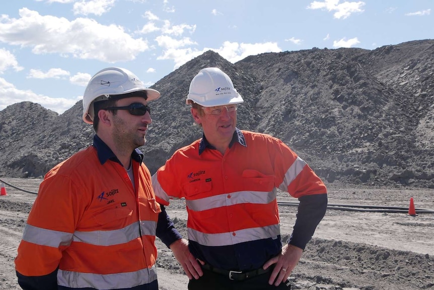 Two men wearing bright orange shirts and hard hats stand in an open-cut mine pit