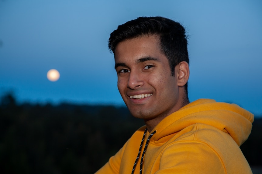 A young man in a yellow jumper smiles at the camera. The moon shines in the sky behind him