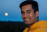 A young man in a yellow jumper smiles at the camera. The moon shines in the sky behind him