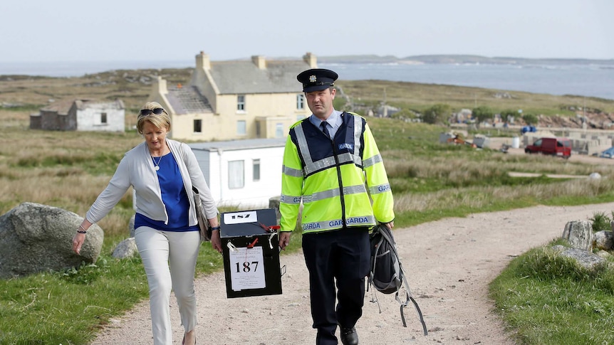 A woman in smart casual dress and a man in a police uniform carry a ballot box along an unpaved road by the sea