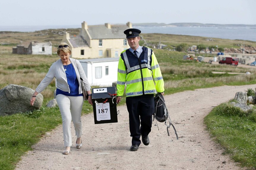 A woman in smart casual dress and a man in a police uniform carry a ballot box along an unpaved road by the sea