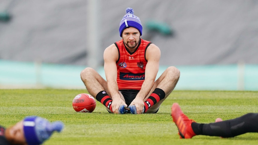 A beanie-wearing AFL footballer sits on the ground at training with his hands touching his feet.