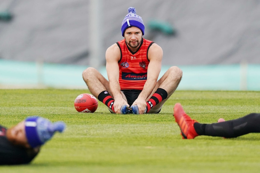A beanie-wearing AFL footballer sits on the ground at training with his hands touching his feet.