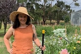 A woman staring at the camera smiling, standing in her garden, flowers are visible behind her right shoulder