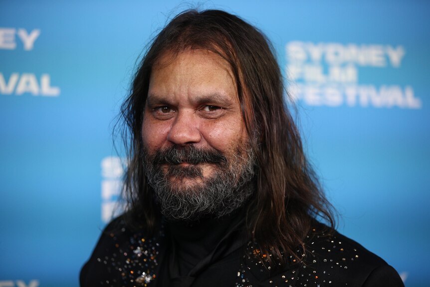 An Indigenous man with long dark hair and a beard smiles for the camera, with a blue film festival backdrop behind him.