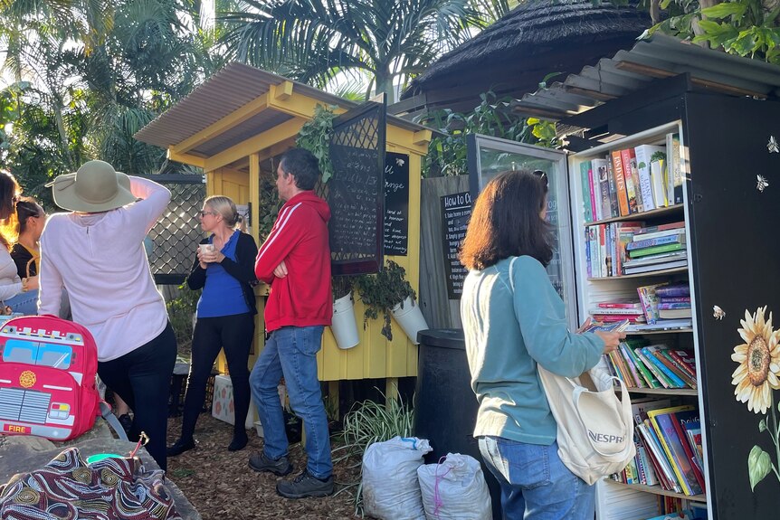 An image of a woman looking at a community library bookshelf and people standing in front of a yellow hutch filled with food