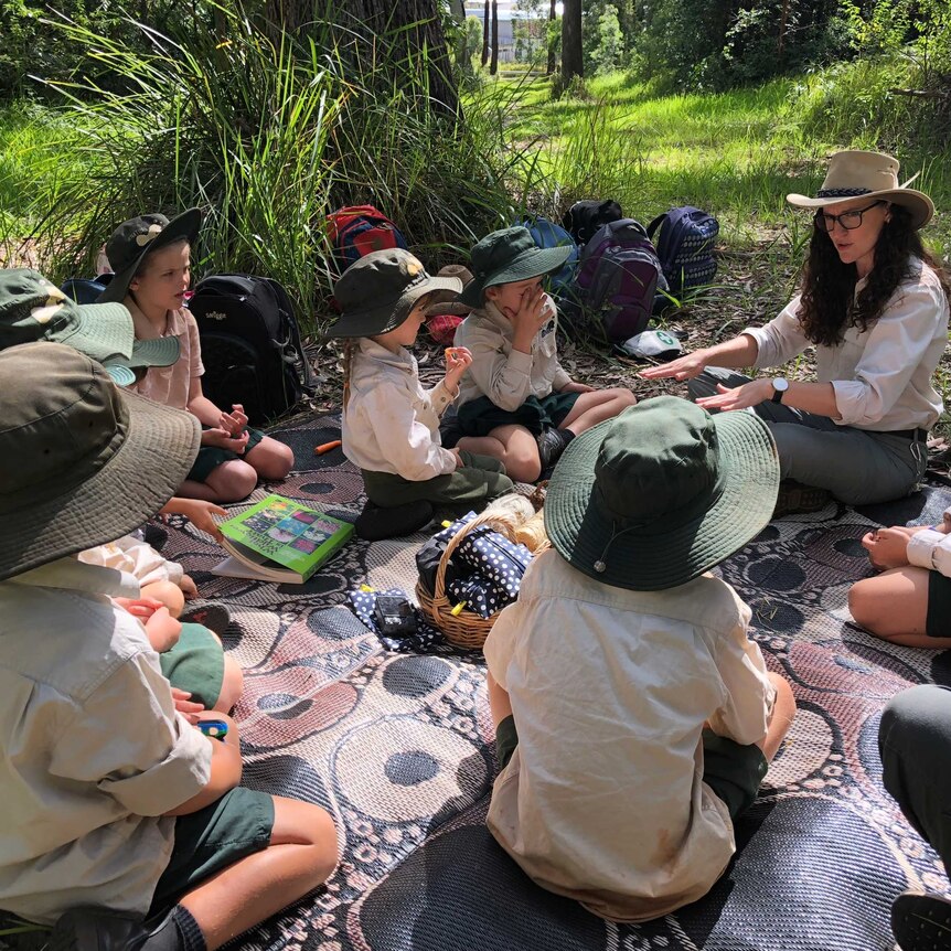 A group of students, wearing hats and uniforms, sit in the sunshine on a large mat in a parkland as they listen to their teacher