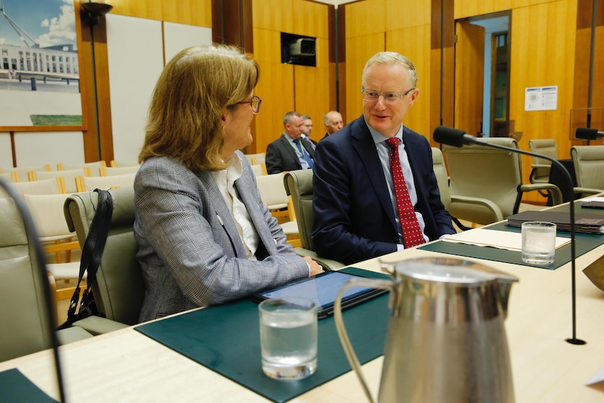 Philip Lowe and Michele Bullock talk and smile while sitting in a committee room at Parliament House