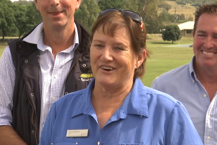 A female politician outdoors at a press conference flanked by two male politicians