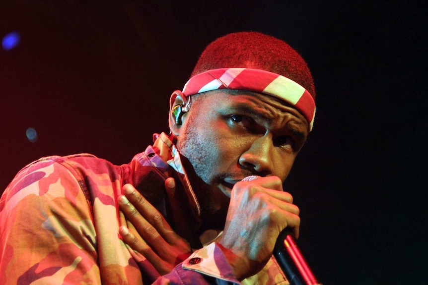 Frank Ocean on stage, singing into a microphone, wearing a red and white headband, with his right hand over his heart