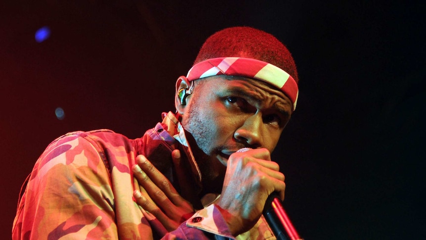 Frank Ocean on stage, singing into a microphone, wearing a red and white headband, with his right hand over his heart