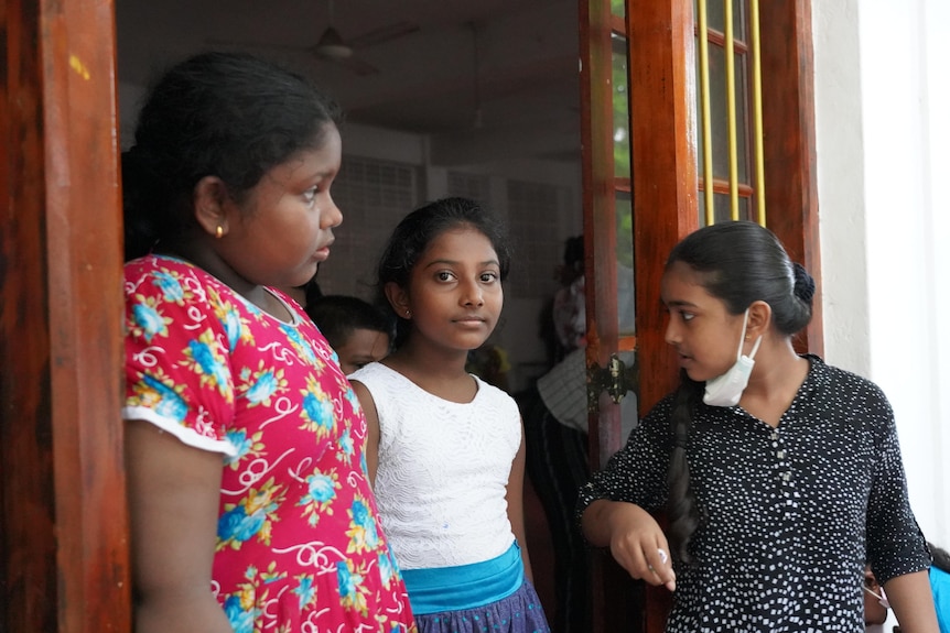 Three pre-teen girls stand in a doorway, one looks towards the camera while her friends look elsewhere