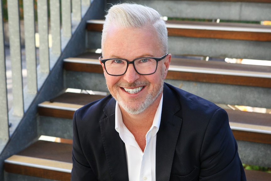 Gary Mortimer wears a white open collared shirt, navy suit jacket and black rimmed glasses and sits on an outdoor staircase