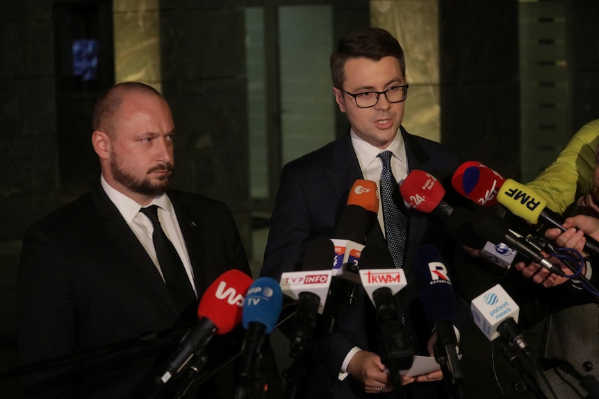 Two men in suits speak into an array of press microphones.