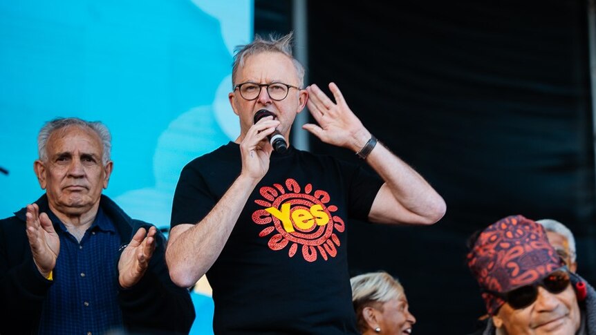 Prime Minister Anthony Albanese – wearing a shirt that says "Yes" – speaks into a microphone while a man applauds behind him.
