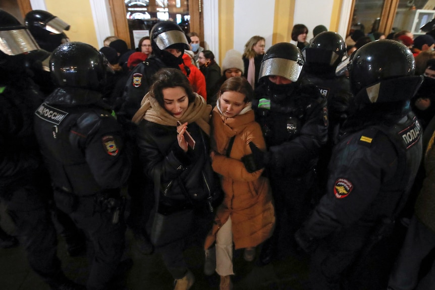 A group of Russian police officers dressed in uniform surroud two women wearing coats.