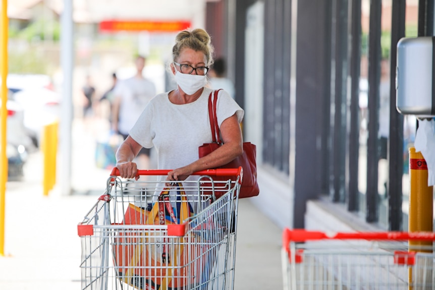 A woman wearing a white shirt and white face mask wheeling a trolley into a supermarket.