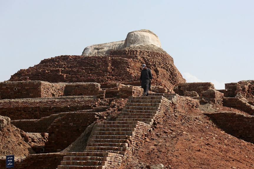 Brick and sand ruins, including multiple flights of steps, rise iout of red dirt as a man walks down. 