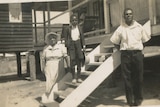 An old photo of an indigenous woman, boy and man, formally dressed,  standing around stairs in front of an old building.