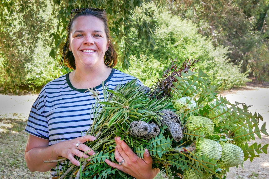 A women smiles at the camera while holding a bunch of freshly picked Australian native flowers. 
