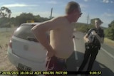 A shirtless man about to be arrested by police on the side of a road.