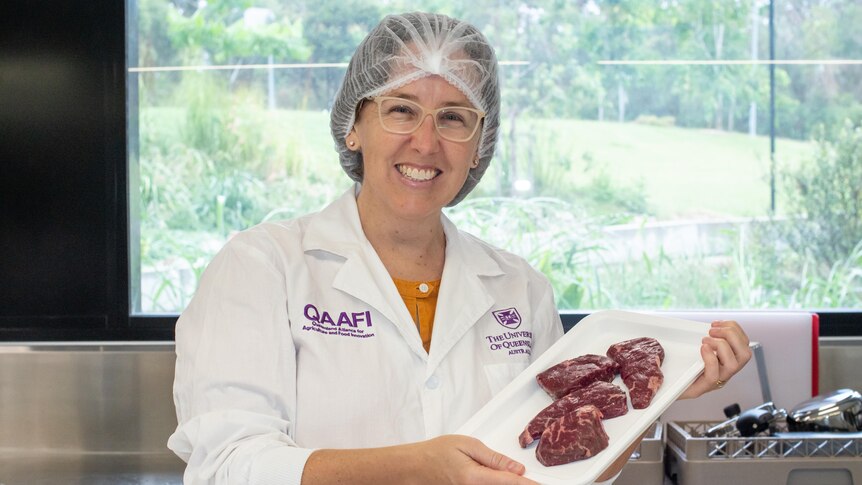 A woman wearing a labcoat and hair net holds a white plate of steaks.