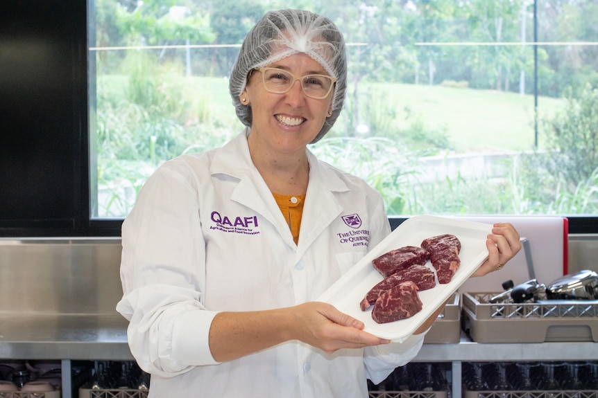 A woman wearing a labcoat and hair net holds a white plate of steaks.