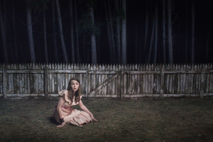 Bethany Whitmore sits on grass with an old wooden fence behind her and a forest beyond the fence.