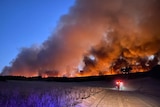 A bushfire burns on a farm paddock at dusk with thick smoke billowing into the air and fire trucks on the ground.