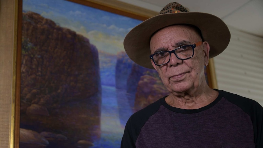 A man in thick rimmed glasses and a hat stands in front of a painting.