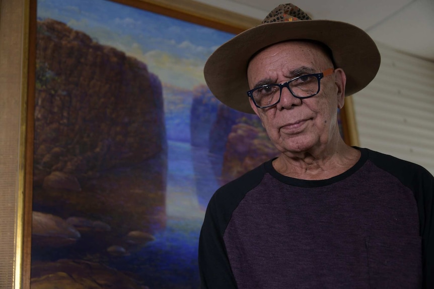 A man in thick rimmed glasses and a hat stands in front of a painting.