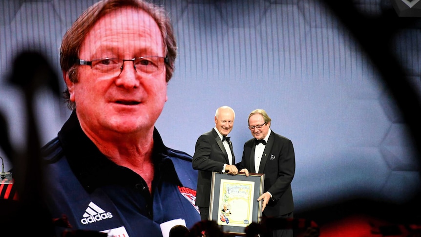 Kevin Sheedy receives a certificate commemorating his Legend status in front of a giant screen showing his face.