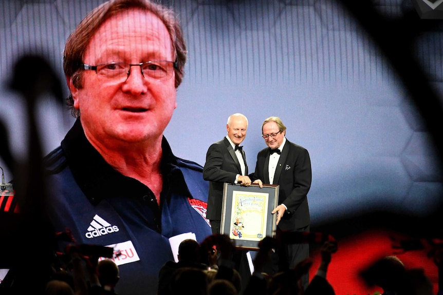 Kevin Sheedy receives a certificate commemorating his Legend status in front of a giant screen showing his face.