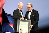 Kevin Sheedy receives a certificate commemorating his Legend status in front of a giant screen showing his face.