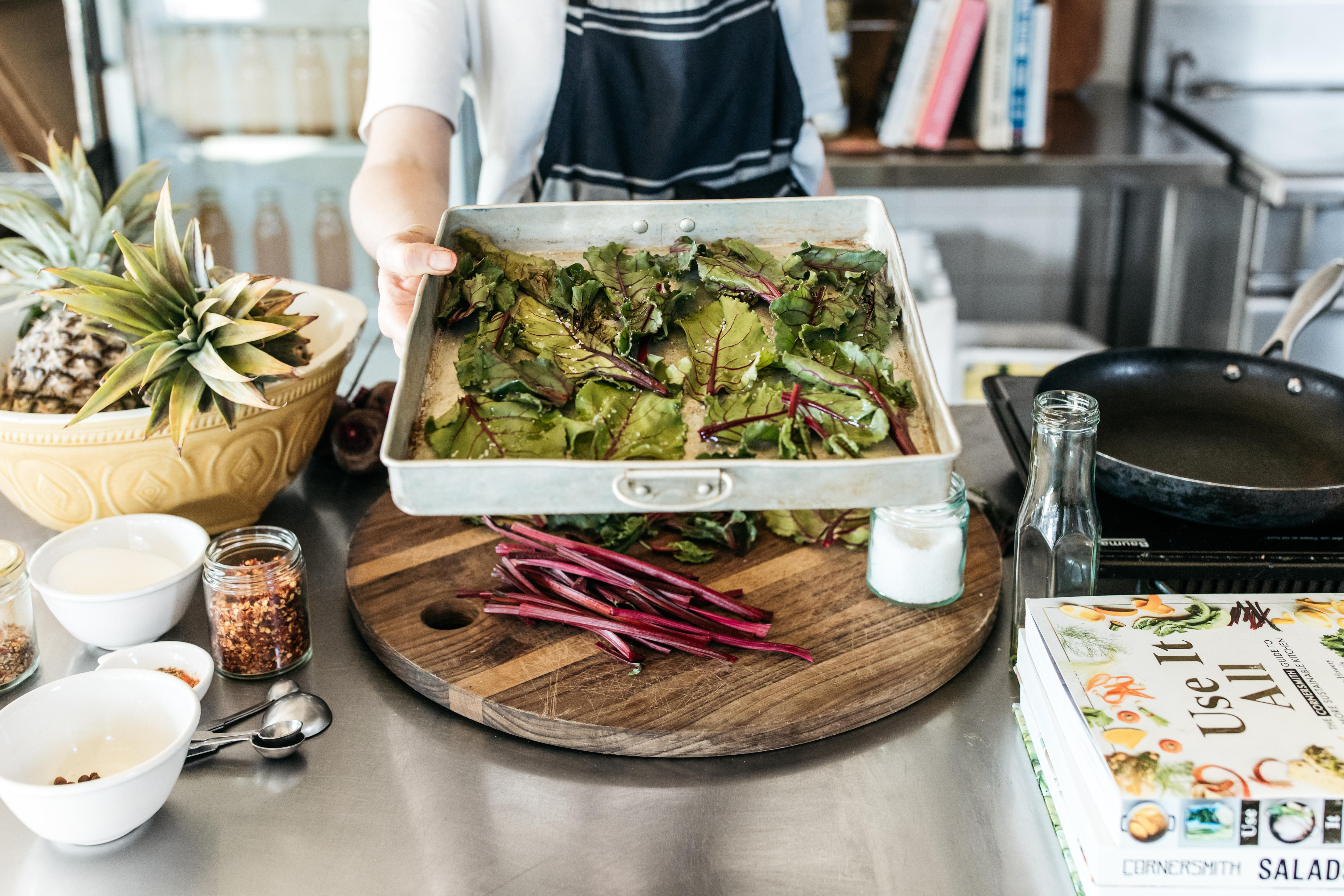A person holds a baking tray out to the camera with beetroot leaves on it while standing in a kitchen.
