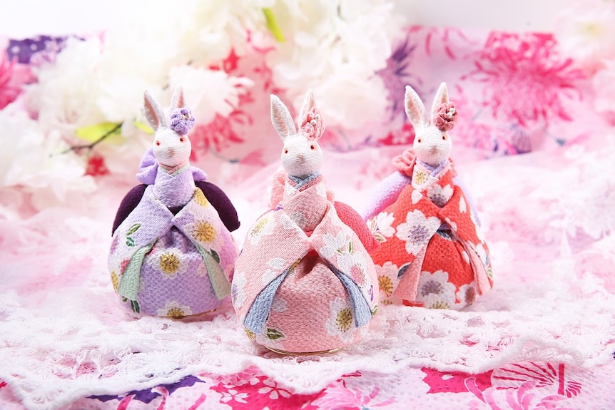 Three small rabbit-like figurines look up  in traditional Japanese dress featuring pink, purple and red flower patterns.