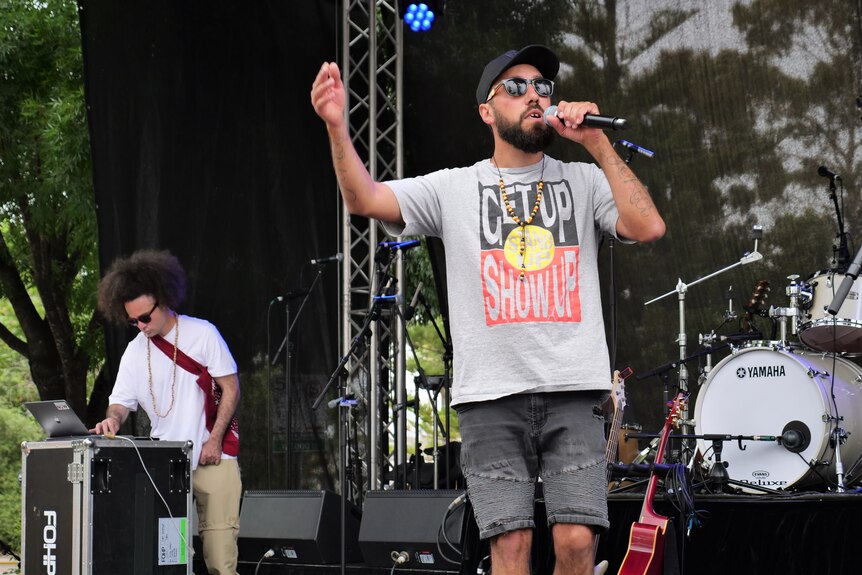 A man holding a mic raps on stage wearing a grey shirt, a cap and sunnies. 