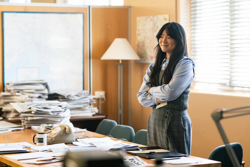 A 30-something Asian Australian woman with curtain bangs stands smiling, with her arms folded in a 1980s-style news office.