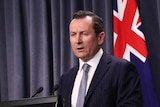 WA Premier Mark McGowan standing at a lectern in front of blue curtains and three flags.