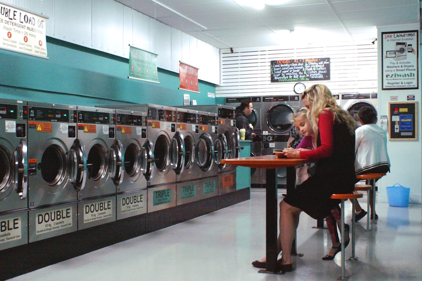 People sitting on stools and standing in front of washing machines at a laundromat.