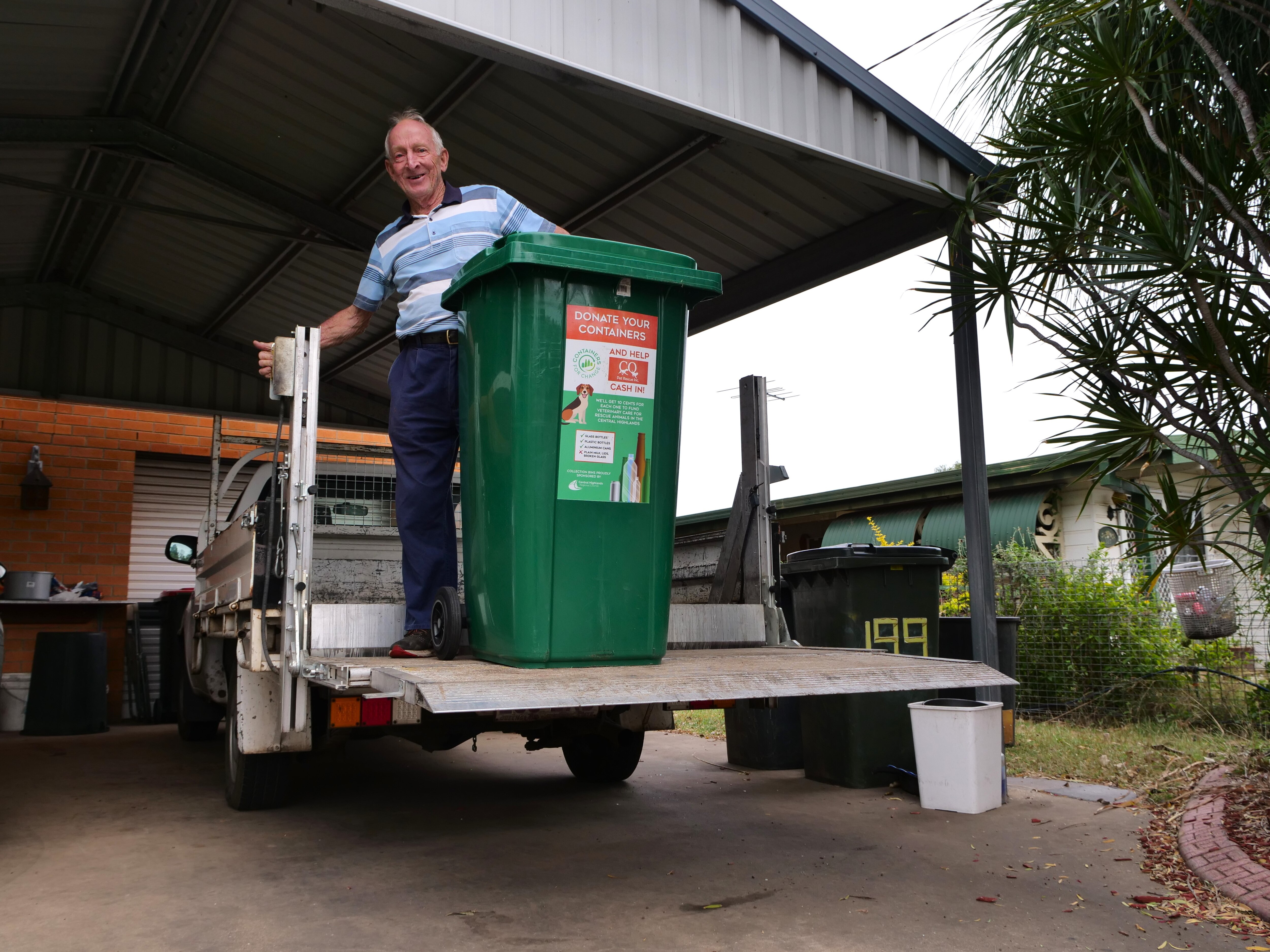 A man standing on a ute tail lifter with a green bin, he is in the car port of his brick home