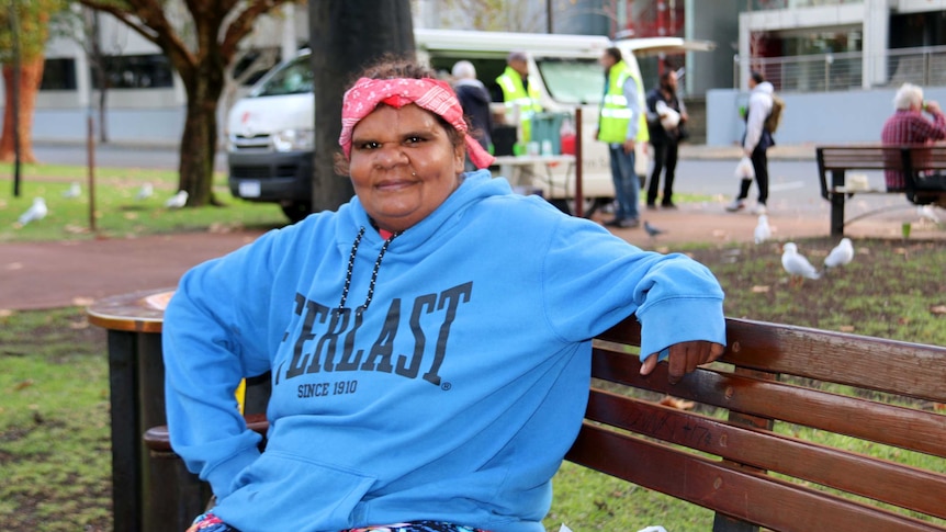 Homeless woman Andrea sitting on a bench with a soup kitchen in the background.