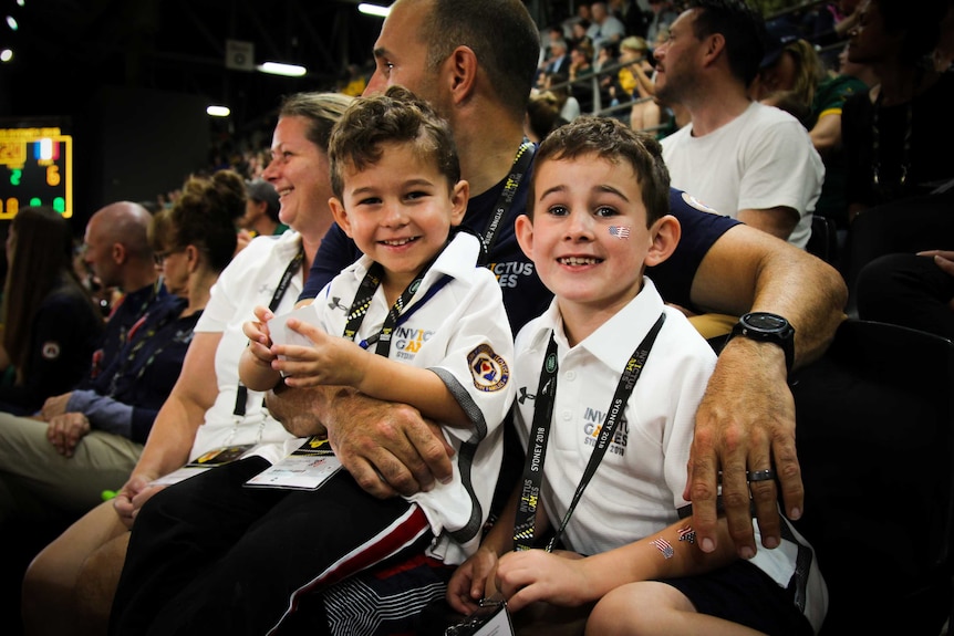 Two young brothers sit on their father's lap in stadium seating.
