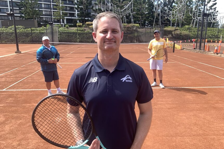 A man wearing a blue polo shirt stands on a tennis court with a racquet in his hand