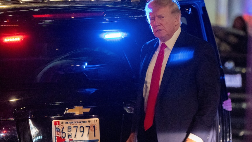 Former US President Donald Trump is pictured in the dark with a black car behind him that has police-like red and blue lights. 