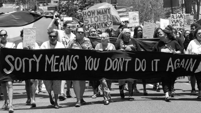 A group of older women walk the streets with a large banner that says 'Sorry means you don't do it again'